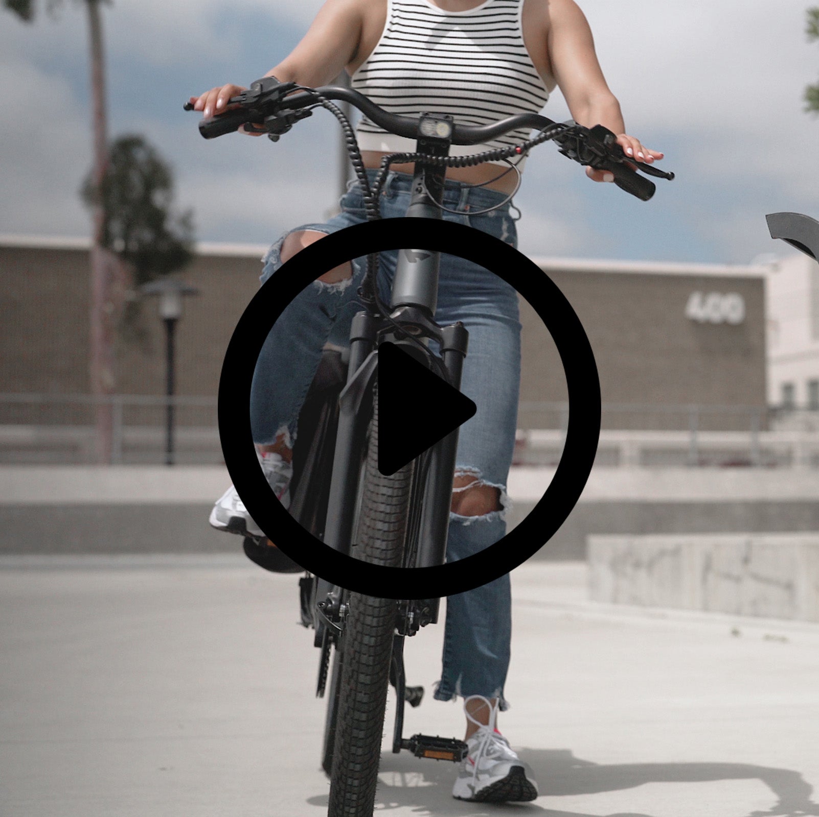 "Front view of a person riding a dark grey electric bike from FavoriteBikes on a concrete surface. The rider is wearing a striped crop top, ripped blue jeans, and white sneakers. The bike features a robust frame, wide tires, and a bright headlight, and the rider appears to be enjoying a casual ride in an urban setting."