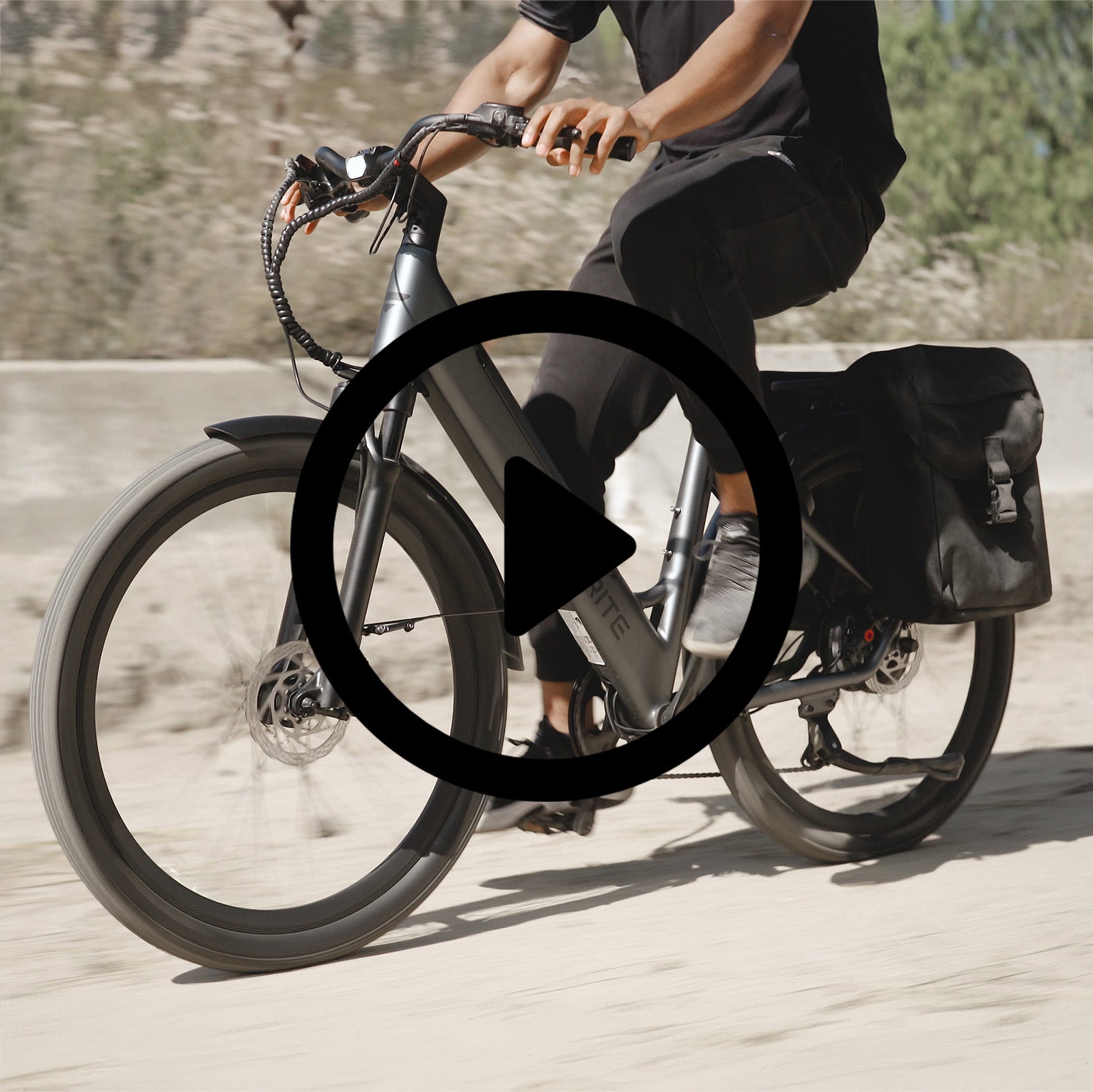 Person riding a sleek, dark grey electric bike from FavoriteBikes on a sandy path, with a black pannier bag attached to the rear. The bike features a modern design with a sturdy frame, hydraulic disc brakes, and a bright headlight. The rider is dressed in black clothing and appears to be enjoying a smooth ride.